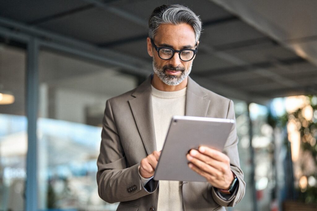 Man in a blazer and glasses reading a tablet while standing in a modern office space
