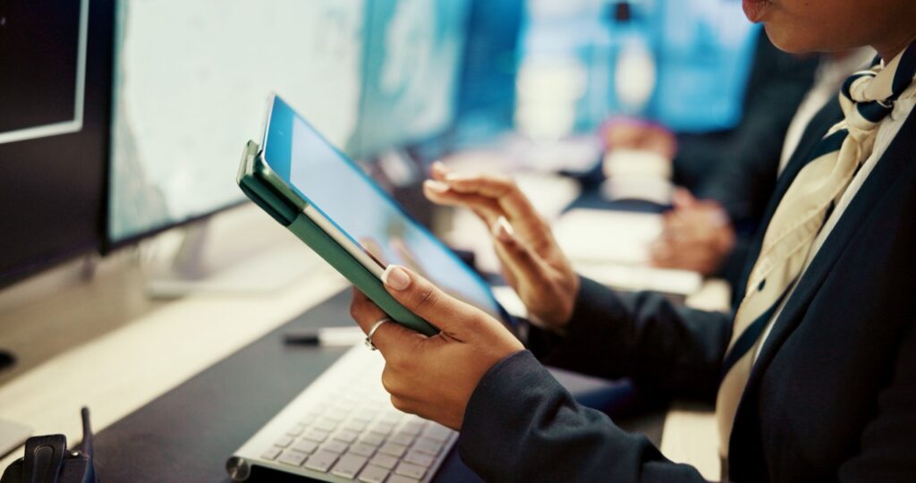 Business professional in a suit reviewing a tablet in an office meeting, with colleagues and computer screens in the background.