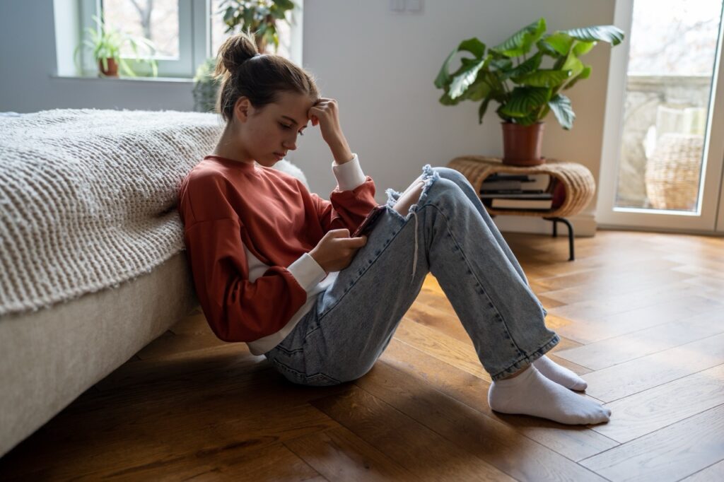 Teenage girl sitting on the floor leaning against a bed, looking down at her phone with her hand on her forehead.