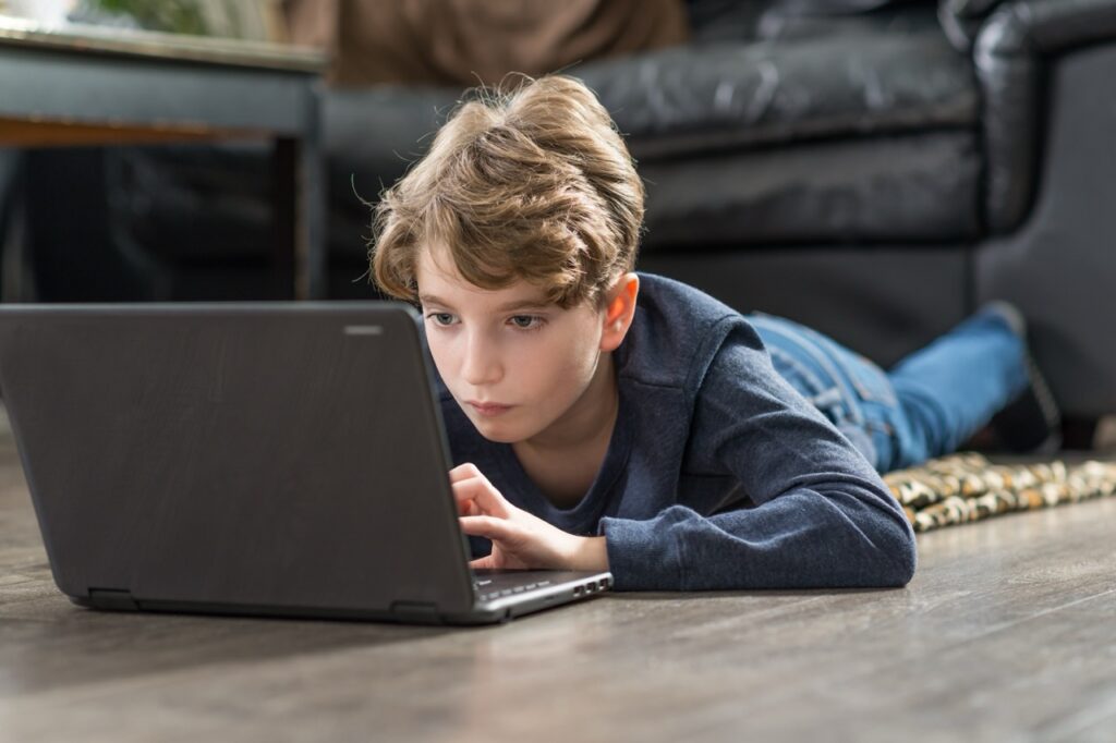 A young boy lies on his stomach on a wooden floor at home, focused intently on a laptop screen in front of him. He rests on his forearms with one hand on the trackpad, wearing a navy jumper and jeans, with a black leather sofa visible in the background.