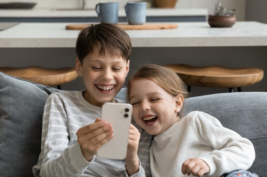 Two young siblings share a smartphone at home, laughing at content on the screen